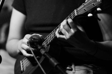 Sahnede ukulele çalan bir müzisyenin ön planda mikrofonuyla yakın çekimi. Ellere ve tellere odaklanan siyah ve beyaz görüntü.