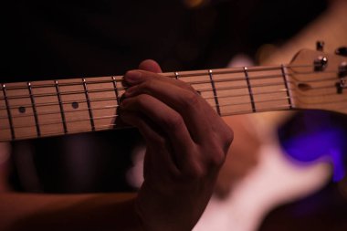 Karanlık bir sahnede gitar çalan bir elin yakın çekimi. Fretboard ve parmaklarına odaklanmış..