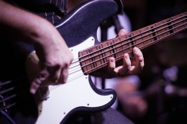 Bas gitar çalan bir elin yakın çekimi, odak noktasında teller ve parmaklar, arka plan bulanık. Bir caz gösterisinin ritmini ve sanatını yakalıyor..