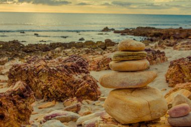 Gün batımında kayalık bir sahilde dengeli taşlar yığını. Arka planda okyanus var. Batan güneşin ılık ışığı kayaların ve denizin dokusunu ve renklerini güçlendirir..
