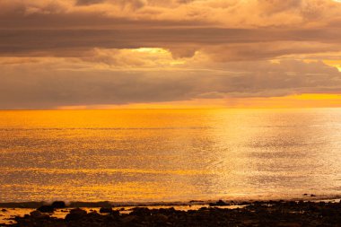 Sakin sulara ve dramatik bulutlu gökyüzüne yansıyan altın ışıkla okyanusun üzerinde sakin bir günbatımı..