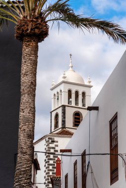 Dar bir sokaktan görünen kubbesi olan beyaz kilise kulesi, bir palmiye ağacı tarafından kısmen gizlenmiş..