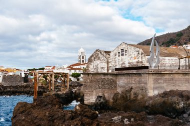 Yıkanmış beton bir yapı ve paslı metal iskelet arka planda güzel bir köy ve kilise bulunan sakin bir okyanusa tepeden bakan karanlık volkanik kayalar üzerinde duruyor. Garachico, Tenerife