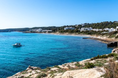 Menorca, İspanya - 11 Mayıs 2024: Açık mavi gökyüzünün altında beyaz binalar ve yeşil tepeler olan bir plajın yakınındaki turkuaz suda bir tekne yüzüyor.