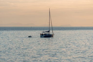 Menorca, İspanya - 11 Mayıs 2024: Gün batımında sakin sularda yüzen bir yelkenli, ufukta görünen uzak bir kıyı şeridi ile.