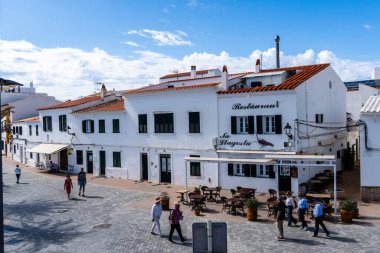 Fornells, İspanya - 12 Mayıs 2024: Kırmızı kiremit çatılı beyaz binalar, insanların Sa Llagosta adlı bir açık hava restoranında gezinip yemek yedikleri güneşli bir plaza..