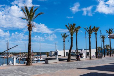 Fornells, İspanya - 12 Mayıs 2024: Palmiye ağaçları güzel bir günde teknelerle dolu sakin bir marinaya bakan güneşli bir liman meydanı hizasında..