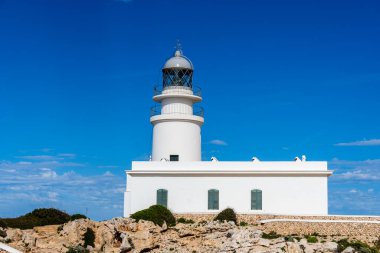 Menorca, İspanya - 12 Mayıs 2024: Kayalık bir kıyı şeridine bakan parlak mavi gökyüzüne karşı heybetli bir beyaz deniz feneri duruyor.