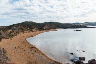 Menorca, İspanya - 12 Mayıs 2024: Sakin deniz ve bulutlu gökyüzünün altında yeşil tepelerle çevrili, kıyıda dinlenen az sayıda insan. Cala Süvarisi
