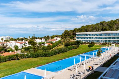 Santo Tomas, İspanya - 12 Mayıs 2024: Beyaz şezlonglu ve şemsiyeli bir tatil köyünde uzun, dikdörtgen bir havuz, beyaz binalar ve mavi bir deniz manzaralı yeşil bir yamaç.
