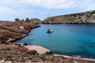 Cala Morell, İspanya - 14 Mayıs 2024: Katamaran tenha bir turkuaz koyda demirledi, engebeli, kırmızımsı kahverengi kayalıklarla çevrili, çoğunlukla açık bir gökyüzü altında.