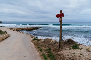 Santo Tomas, İspanya - 15 Mayıs 2024: Taş küreli bir kıyı yolu bulutlu bir gökyüzü altında sakin bir plaj sahnesine çıkar..