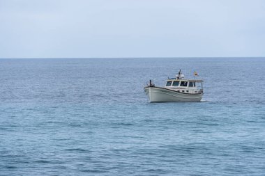 Mao, İspanya - 15 Mayıs 2024: Soluk bir gökyüzünün altında sakin mavi bir denizde beyaz bir motorlu tekne yüzüyor.