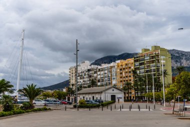 Denia, İspanya - 21 Eylül 2024: Apartman binaları ve palmiye ağaçları bulutlu bir gökyüzünün altında arka planda dağlar bulunan bir şehir caddesi hizasında.