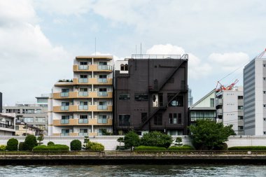 Bulutlu bir gökyüzü altında çeşitli modern mimari sergilenen bir dizi bina nehir kıyısına dizilmiştir..