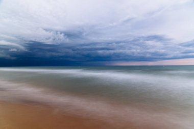 Dalgaların ritmik hareketlerini içeren sakin bir deniz manzarası kasvetli, bulutlarla dolu bir gökyüzünün altında kumlu sahile çarpıyor. Aşağıdan kes