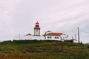 Portekiz 'in batısındaki Cabo da Roca' daki deniz feneri bulutlu bir gökyüzünün altındaki tepede duruyor. Beyaz binaların terakotta çatıları vardır..