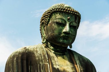 Kamakura, Japonya - 5 Ağustos 2024: Kamakura, Japonya 'daki Büyük Buda heykelinin yakın çekimi, yıpranmış bronz dokusunu açık mavi gökyüzüne karşı sergiliyor.