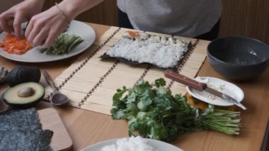 Kadınlar taze malzemelerle suşi yapıyorlar. Tahta bir masada avokado, nori, pilav ve sebze gibi.