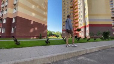 Genç bir birey, arka planda modern binaların yer aldığı sakin bir şehir parkında geziniyor.