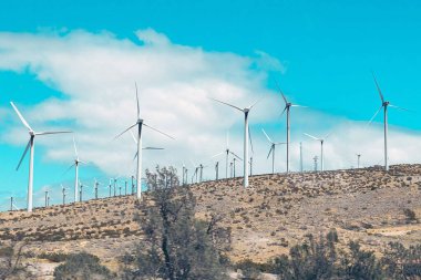 Kuru, tepelik bir arazide, dağınık beyaz bulutlu, açık mavi bir gökyüzünün altında yer alan rüzgar tarlası.