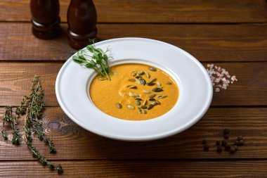 Kabak tohumları ve taze otlarla süslenmiş kremalı balkabağı çorbasıyla dolu beyaz bir kase, ahşap bir masada duruyor.