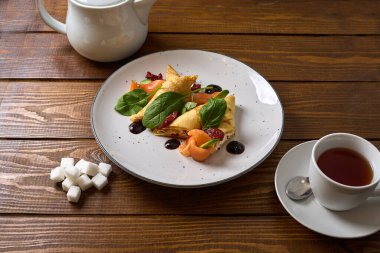 Tütsülenmiş somon, ıspanak ve krem peynirle dolu ince krepler, bir fincan çay ve ahşap bir masada çaydanlıkla servis edilir.