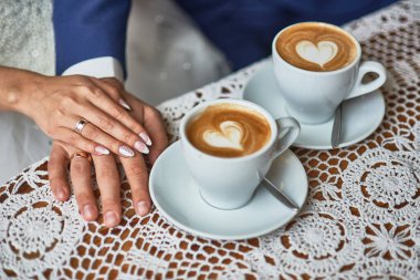 Yeni evlilerin ellerinin ve dantel masa örtüsünün üzerindeki latte sanatının büyüleyici görüntüsü, aşkı simgeliyor, iki fincan, romantik kalp şekli, kahve, birlik, özel bir gün konsepti.