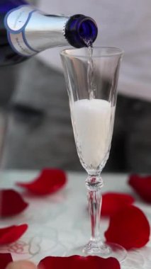 Romantik kutlama konsepti olarak etrafa saçılmış gül yapraklarıyla kristal flüte köpüklü şarap dökmek.