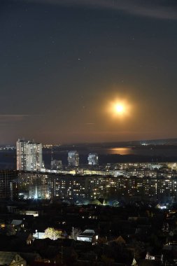 Bright Moon Shines on Cityscape, River Reflection in Urban Scene, Starry Night Sky, Evening Ambiance, City Lights, Celestial Mood Concept clipart