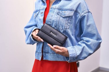 Stylish black wallet held by woman in denim jacket and red dress, featuring modern accessory and fashion ensemble concept clipart