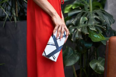 Kırmızı elbiseli, şık debriyaj çantalı sofistike bir bayan, yemyeşil bitki arka planına karşı kurulmuş, tarz aksesuar konsepti