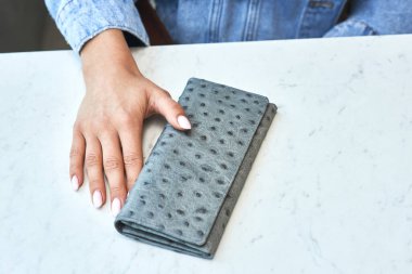 Mermer yüzeyde şık gri deri cüzdan, zarif bir kadının eline manikürlü tırnaklarla dokunmak, aksesuar moda konsepti.