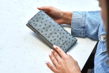 Mermer masa üzerinde devekuşu desenli zarif kadın elleri tarafından tutulan gri deri cüzdan moda ve tarz kavramını vurguluyor.