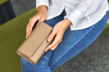 Bir kadın kucağında bej bir cüzdan tutuyor, günlük kot pantolon ve beyaz gömlek giyiyor, manikür ve moda aksesuarları ile düzgün eller sergiliyor.
