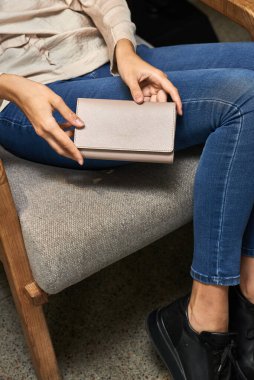 Sandalyede oturan kadının bej cüzdanı. Kucağında dinleniyor. Kıyafet detaylarını sergiliyor.