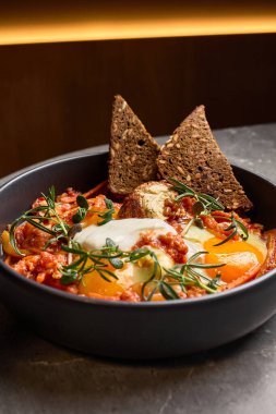 Lezzetli Shakshuka 'da haşlanmış yumurtalar, domatesler, siyah bir kasede otlar, gevrek ekmek ve kırsal kahvaltı konseptiyle tamamlanmış.