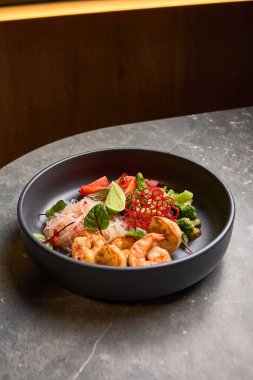 Lezzetli karides yanında taze limon, renkli sebzeler ve gri taş bir masada şık siyah bir kase içinde pirinç eriştesi, Asya mutfağı kavramı.