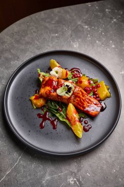 Izgara somon, canlı turunçgil ve siyah bir tabakta nar garnitürle servis edilir. Zarif bir yemek sunumunu temsil eder.