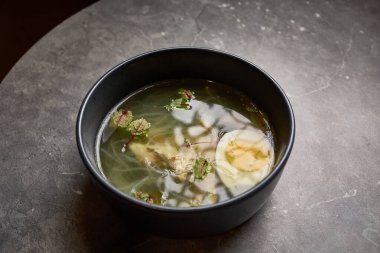 Yumurtalı, otlu ve erişteli berrak çorba, gri mermer masanın üzerinde, rahatlatıcı ve besleyici cazibesini, mutfak stili kavramını vurguluyor.