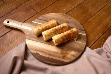 Üç gevrek, altın sarısı kızarmış börek, ahşap bir tahta kesme tahtasına yerleştirilmiş, sıcak ahşap bir masa arkasına kurulmuş..