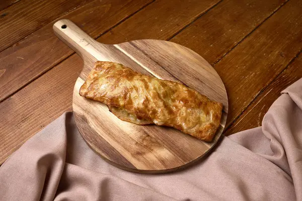Kırsal bir ahşap masanın üzerindeki ahşap kesme tahtasının üzerinde duran, çıtır çıtır altın hamurlu, taze pişmiş dikdörtgen puf böreği..