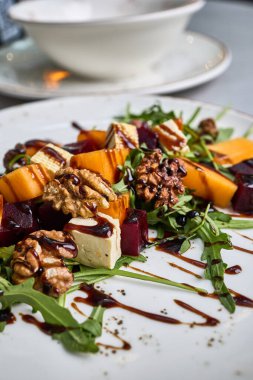 Beyaz bir tabakta kızarmış pancar, hurma, ceviz, roka, brie peyniri ve balzamik jöleli gurme salatasının detaylı bir görüntüsü..