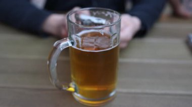 Kehribar birayla dolu bir bira bardağının yakın çekimi, arka planda bulanık bir insanla ahşap bir masaya kurulmuş..