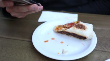 Izgara burrito soslu beyaz bir tabakta kısmen yenir, masada arka planda akıllı telefon kullanan biri..