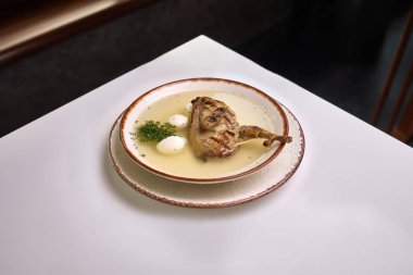 İçinde ızgara bıldırcın bacağı, bıldırcın yumurtası ve kırsal seramik bir tabakta servis edilen taze otlar bulunan bir kase temiz bıldırcın çorbası..