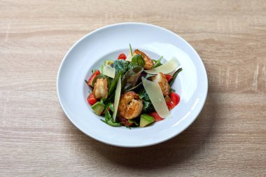 Izgara karides, avokado küpleri, kiraz domatesleri, yapraklı yeşiller ve beyaz tabakta servis edilen parmesan talaşı ile renkli salata..