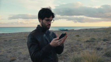 Gözlüklü ve ceketli adam okyanus kıyısındaki sahilde akıllı telefonu kullanıyor.