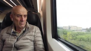 Kulaklıkla müzik dinleyen yaşlı kel adam trenin camından dışarı bakıyor..