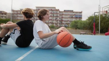 İki genç erkek dışarıda mavi alanda basketbol oynuyorlar..